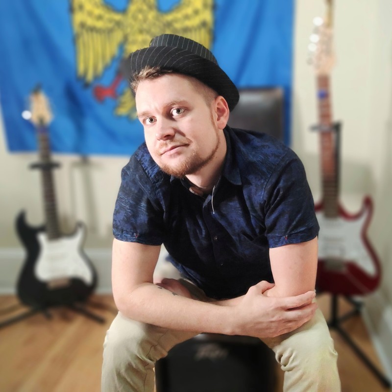 Young man in a black hat and blue patterned shirt sitting on a chair, with arms crossed, in front of a colorful flag and electric guitars in the background.